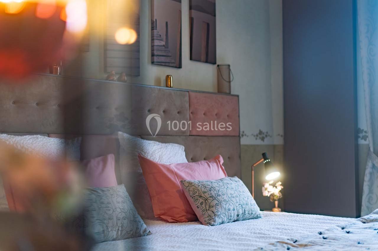 Lit double avec oreillers roses et motifs, éclairé par une lampe de chevet dans une chambre au décor chaleureux.