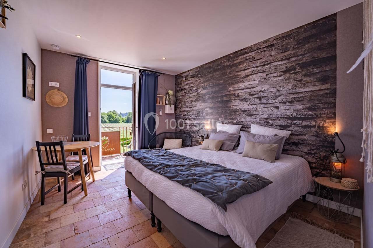 Chambre lumineuse avec lit double, table et chaises, décor rustique et accès à un balcon avec vue sur la nature.