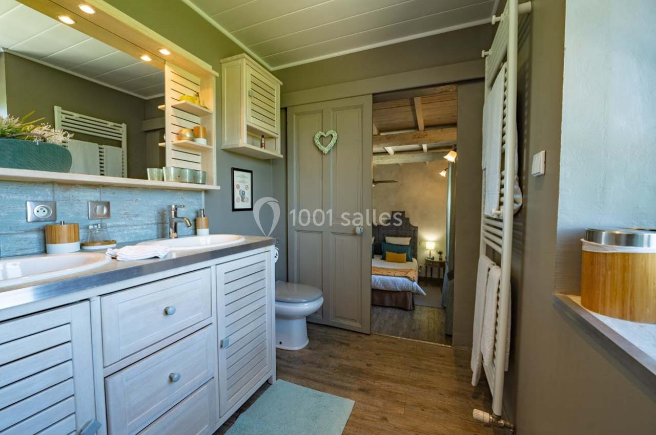 Salle de bain lumineuse avec meuble en bois clair, miroir éclairé, et vue sur une chambre à travers une porte ouverte.