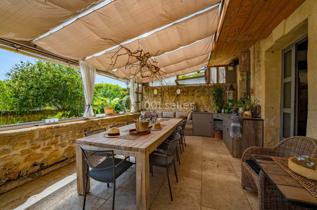 Terrasse extérieure avec table en bois, chaises, plantes et pergola ombragée, entourée de murs en pierre.