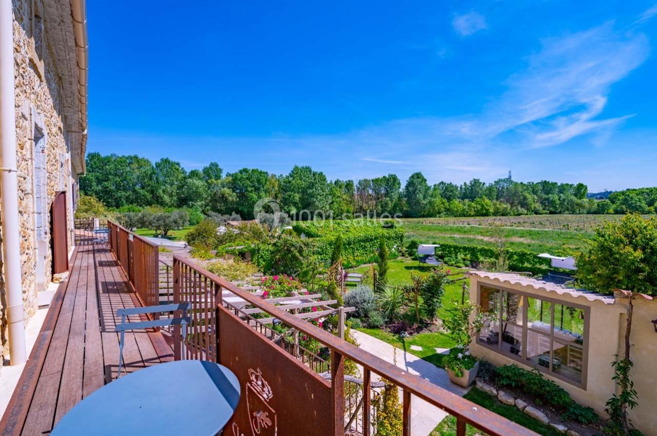 Vue depuis une terrasse en bois donnant sur un jardin verdoyant et des champs sous un ciel bleu dégagé.