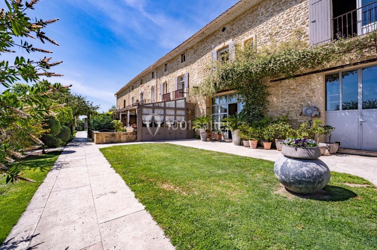 Façade en pierre d'une maison avec jardin verdoyant, pots de fleurs et ciel bleu en arrière-plan.