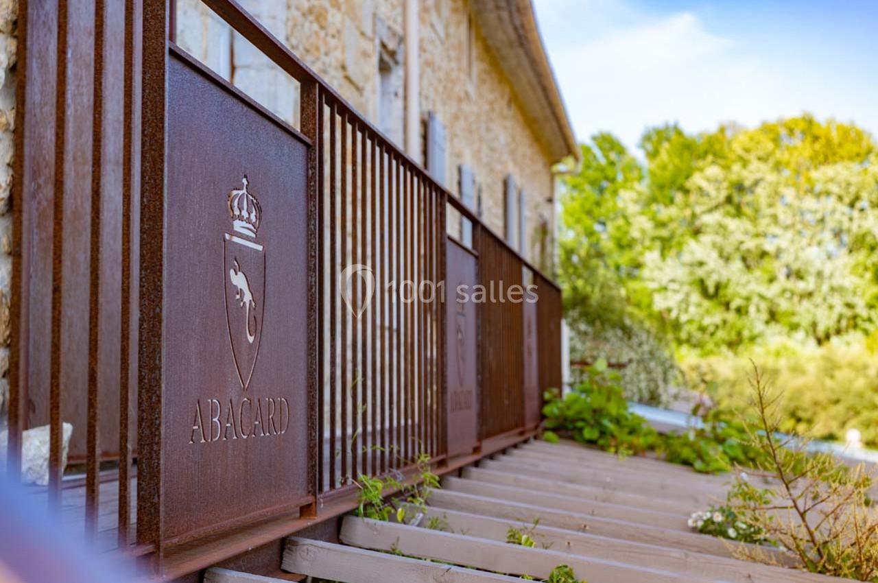 Clôture en métal rouillé avec un logo gravé, longeant un bâtiment en pierre entouré de verdure.
