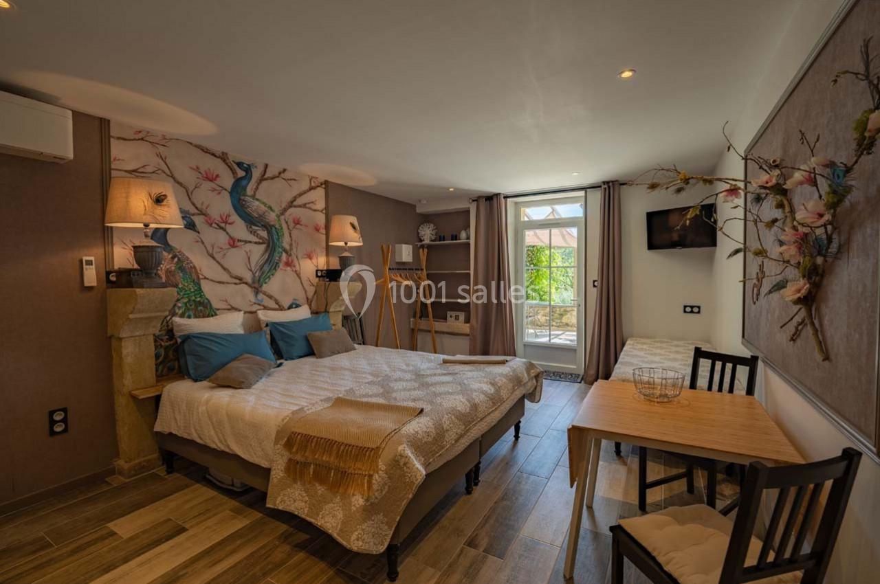 Chambre lumineuse avec lit double, décoration murale colorée, bureau, table, et accès à une terrasse extérieure.