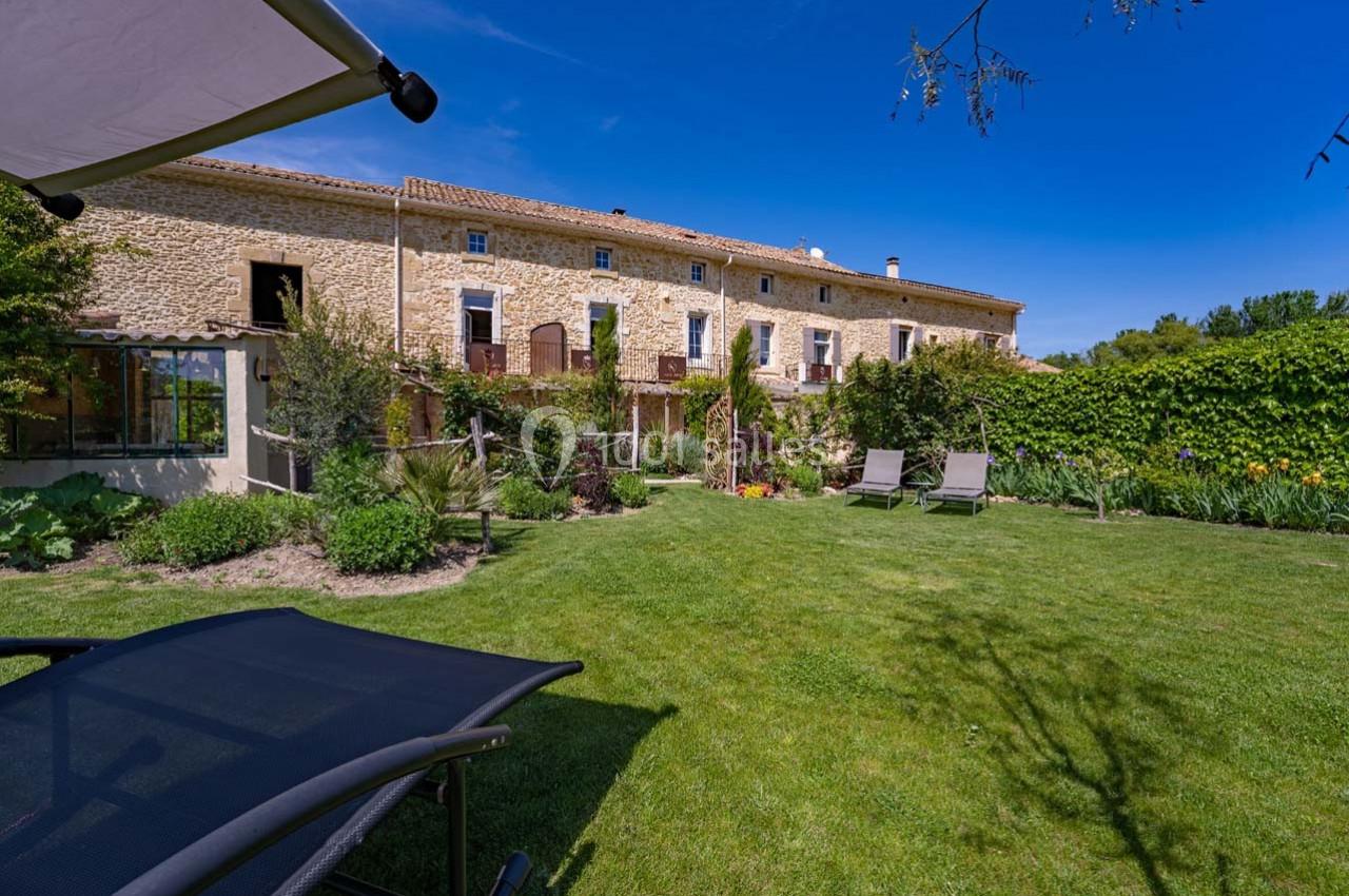 Façade en pierre d'une maison avec jardin verdoyant, chaises longues et parasol sous un ciel bleu.