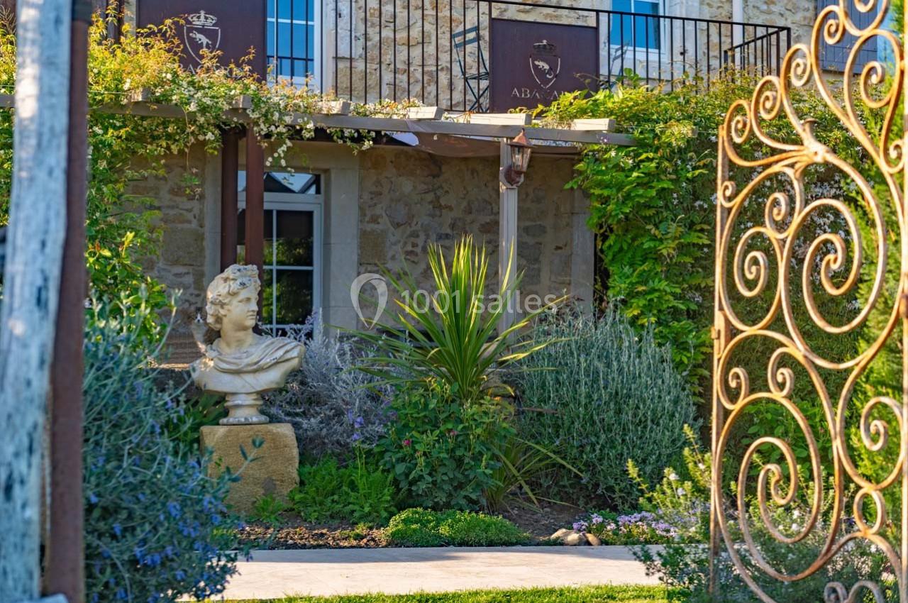 Jardin fleuri avec une statue en pierre, devant une façade en pierre ornée de plantes grimpantes et une porte en fer forgé.