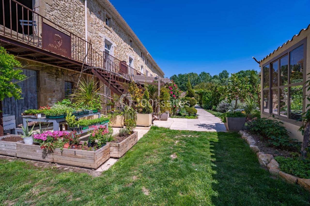 Cour d'une maison en pierre avec jardin fleuri, terrasse en bois et vue sur un chemin bordé de verdure.