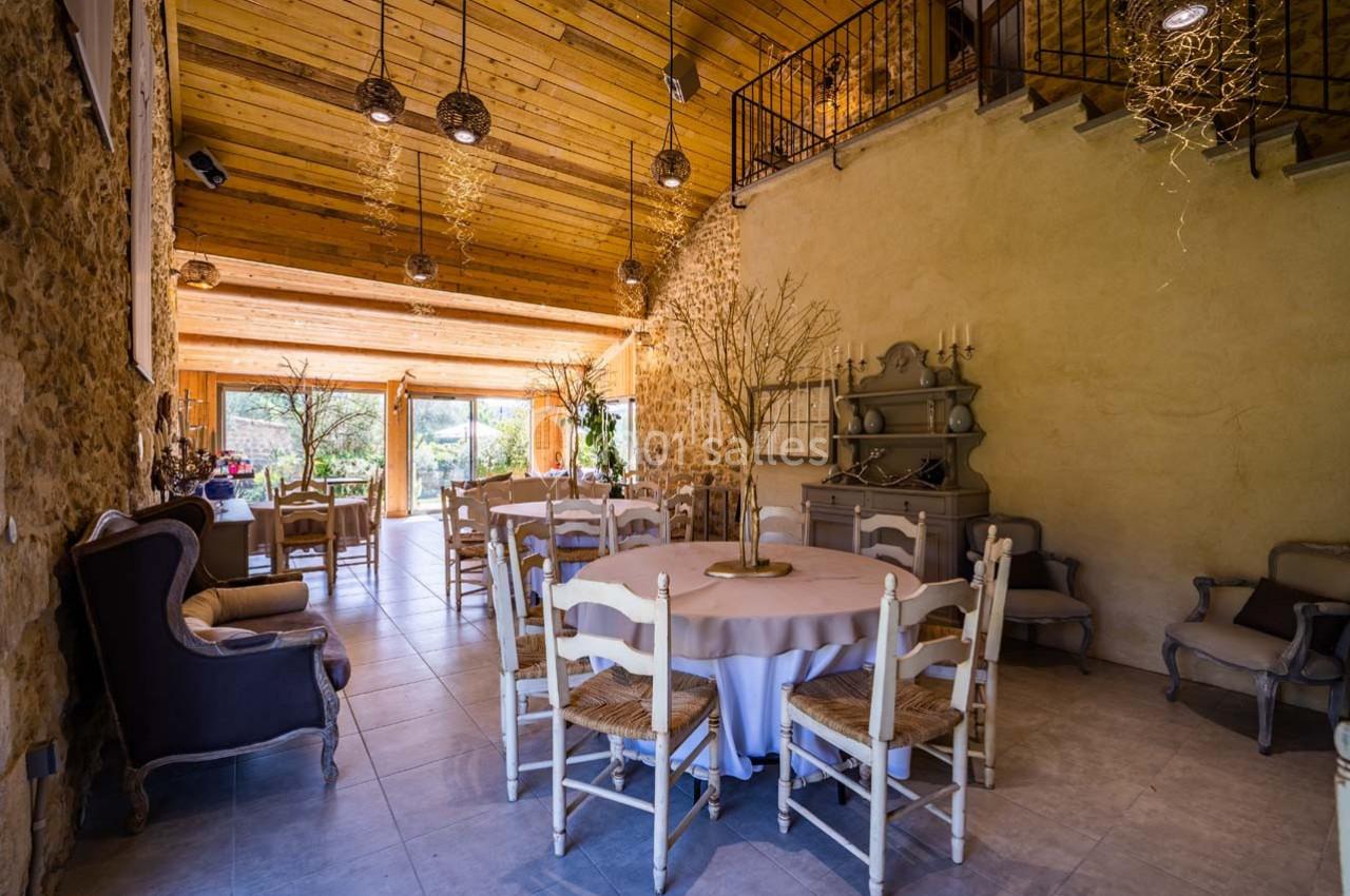 Salle lumineuse avec murs en pierre, plafond en bois, tables rondes dressées et grandes baies vitrées donnant sur un jardin.