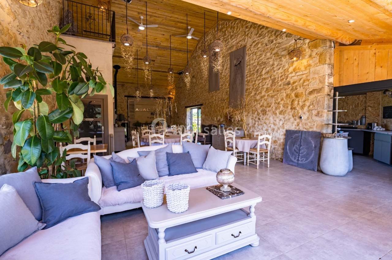 Salle lumineuse avec murs en pierre, plafond en bois, canapés blancs, table basse et chaises autour de tables.