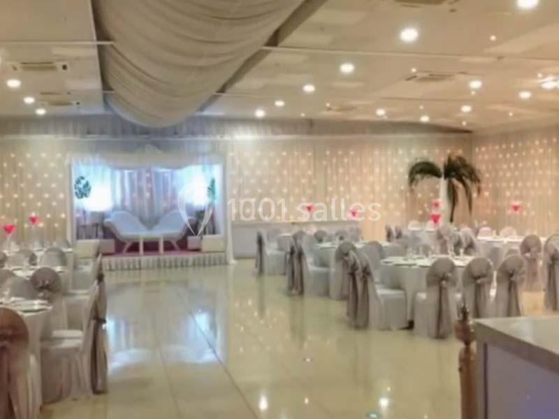 Salle de réception élégante avec tables dressées, chaises décorées et une estrade ornée de rideaux lumineux.