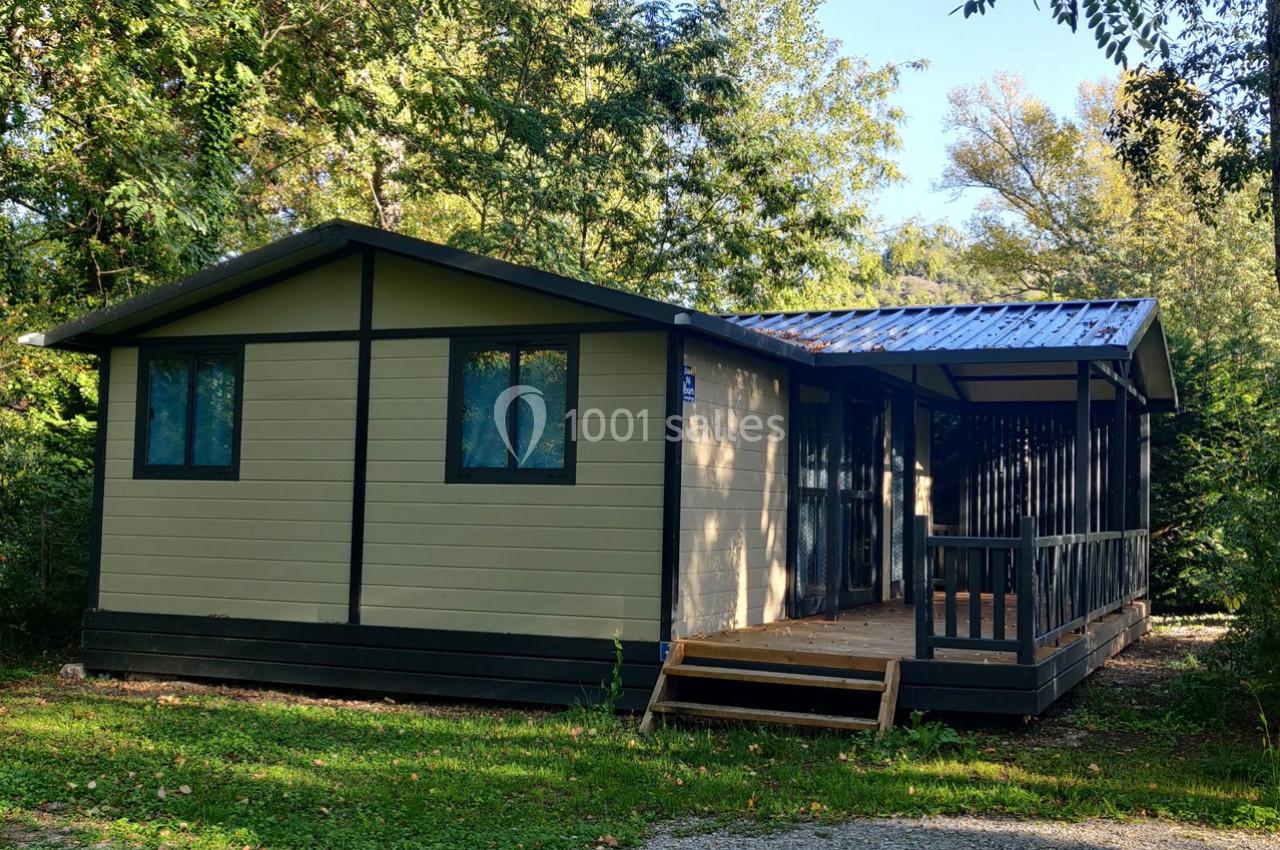 Chalet en bois beige avec terrasse couverte, entouré de végétation et situé sur une pelouse.