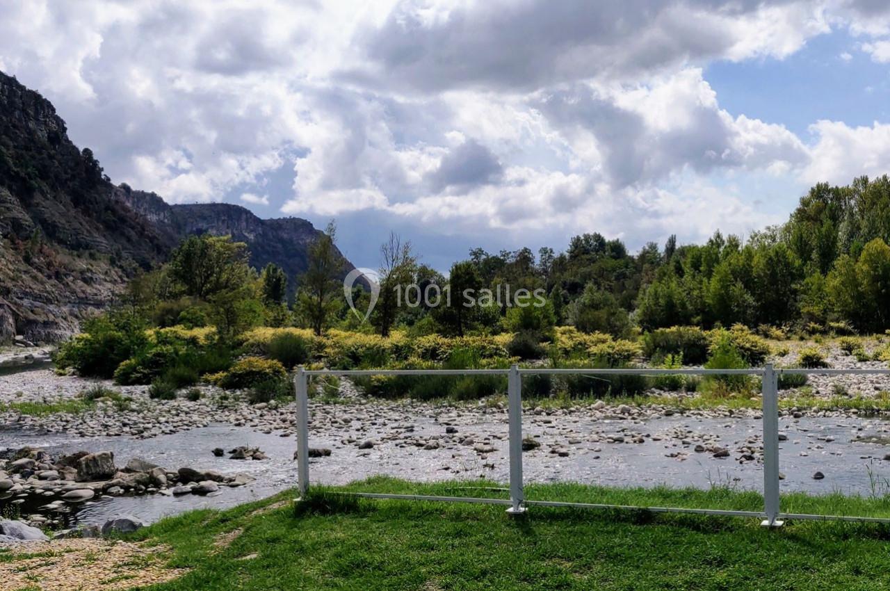 Rivière bordée de végétation et de montagnes sous un ciel partiellement nuageux, vue depuis une barrière blanche.