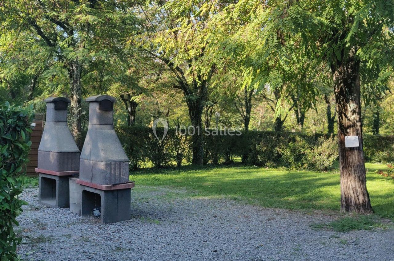Deux barbecues en pierre sur un sol gravillonné, entourés d'arbres et de verdure dans un espace extérieur.