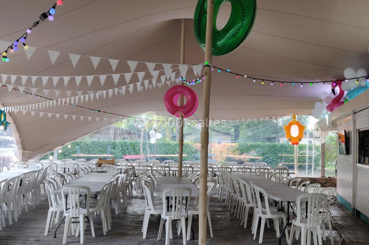 Intérieur d'une grande tente décorée avec des guirlandes lumineuses, des bouées colorées et des tables entourées de chaises…