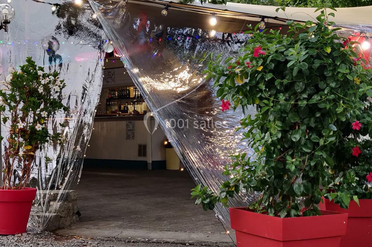 Entrée d'un espace extérieur couvert avec des plantes en pots rouges et une bâche transparente laissant entrevoir un bar.