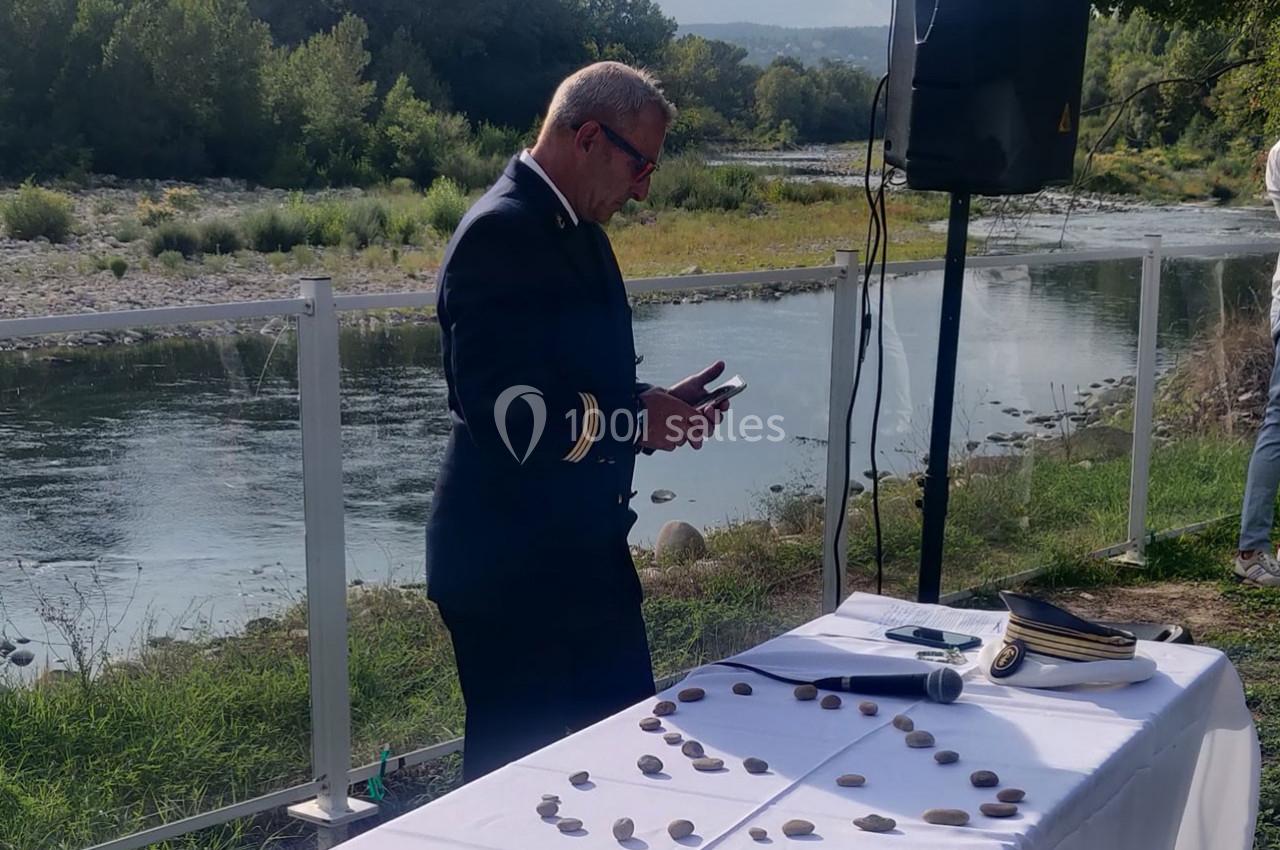 Un homme en uniforme lit un document près d'une table décorée de galets, au bord d'une rivière.