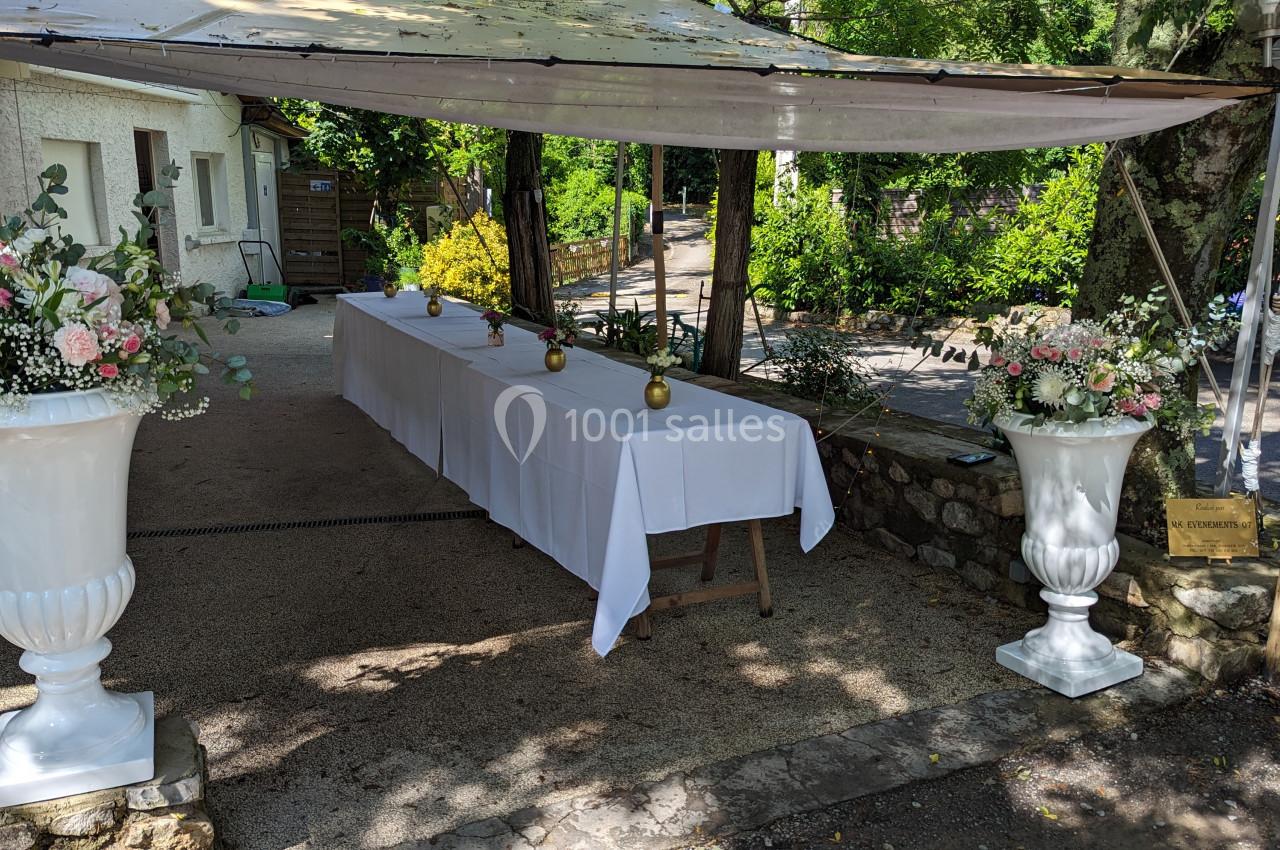 Table décorée avec une nappe blanche sous un auvent, entourée de grands vases fleuris dans un cadre extérieur verdoyant.