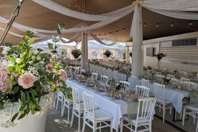 Salle de réception en extérieur décorée pour un événement, avec tables dressées, fleurs et voilages suspendus.