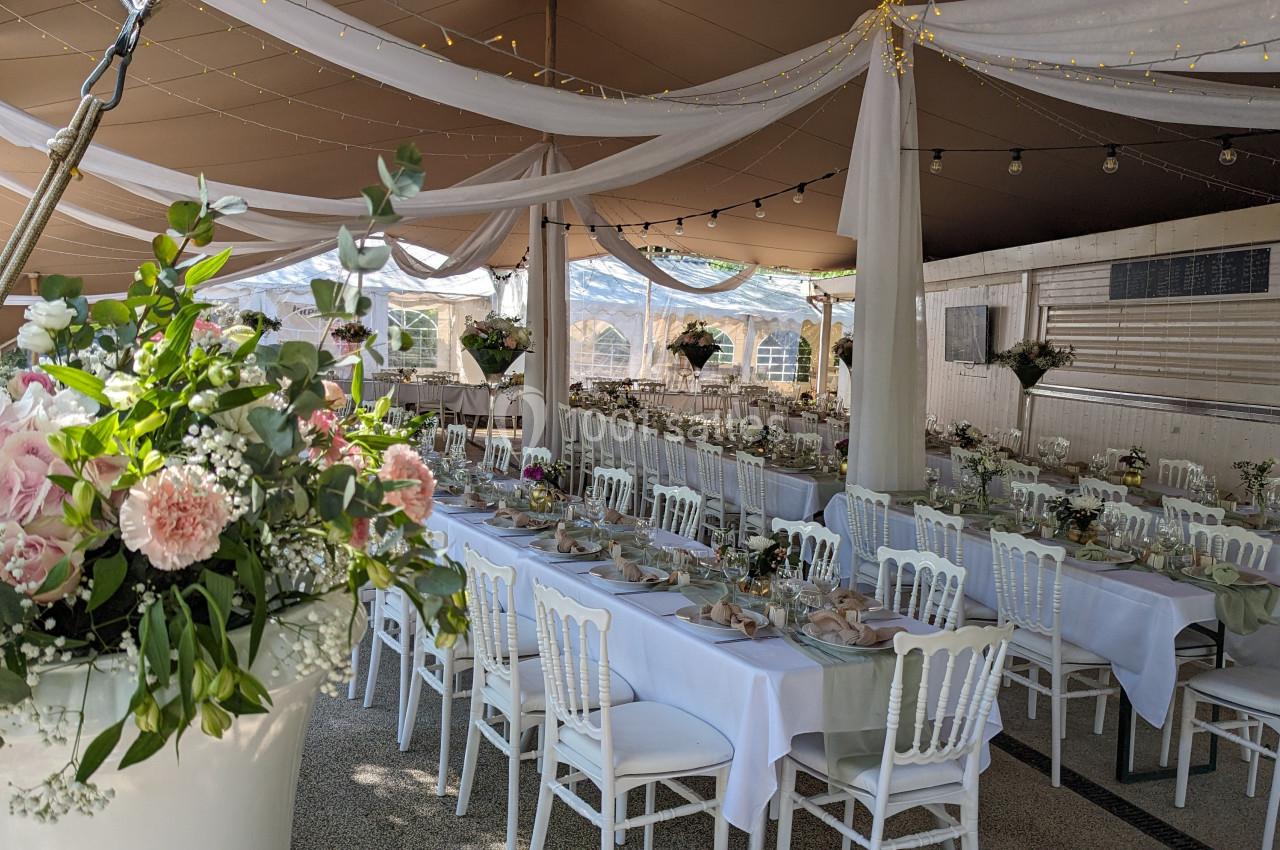 Salle de réception décorée avec des tables dressées, des chaises blanches et des fleurs, sous un chapiteau élégant.