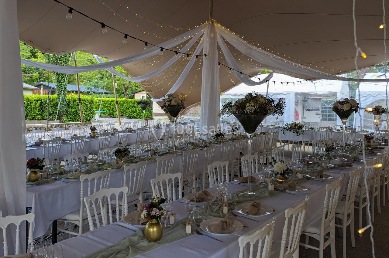 Salle de réception décorée pour un événement, avec longues tables dressées, chaises blanches et guirlandes lumineuses.