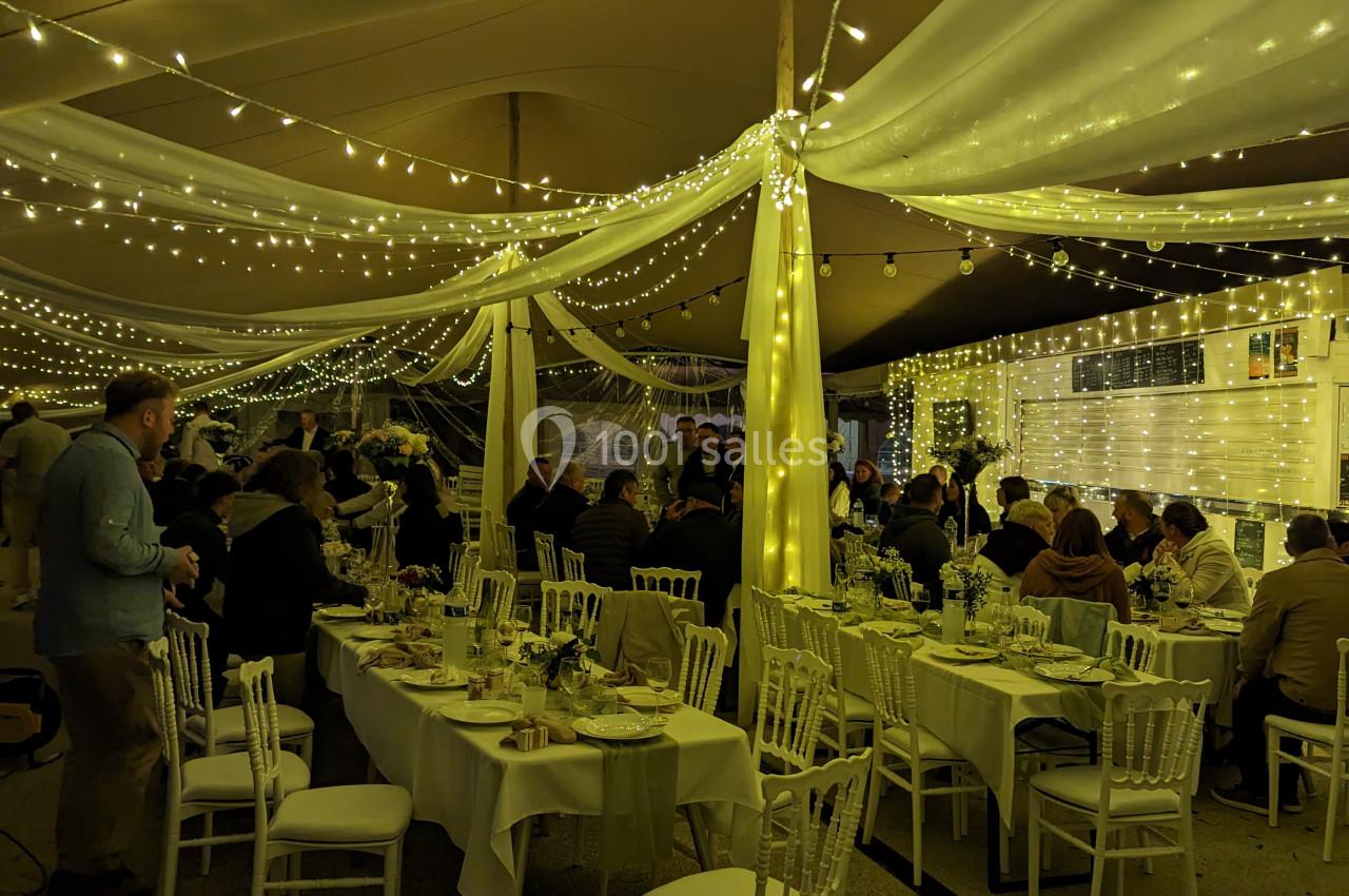 Salle de réception décorée avec des guirlandes lumineuses, tables dressées et invités assis ou debout.