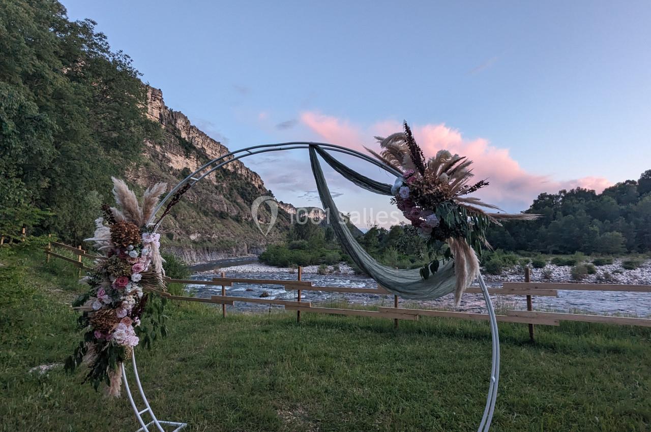 Arche circulaire décorée de fleurs et de tissus, installée dans un paysage naturel près d'une rivière et de collines.