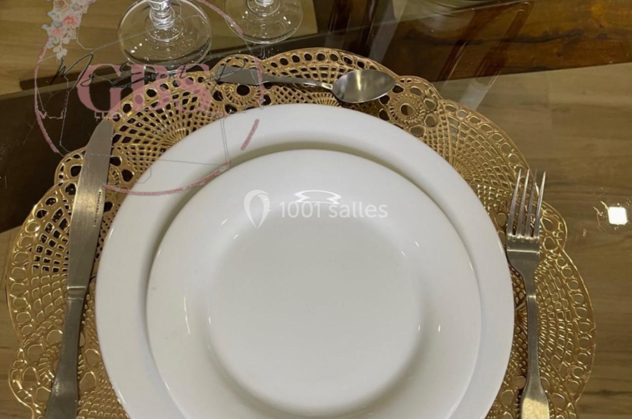 Table dressée avec une assiette blanche sur un napperon doré, couverts en métal et verres en cristal.