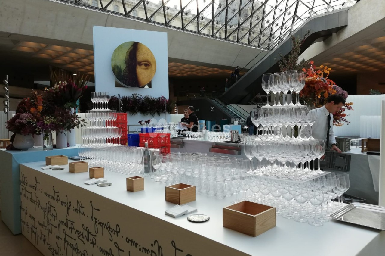 Table dressée avec des verres empilés en pyramide, décor floral et personnel en arrière-plan dans un espace moderne.
