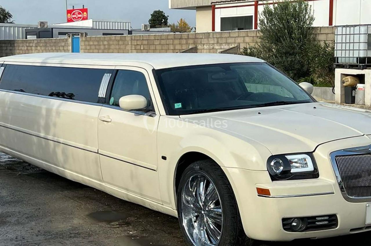 Limousine blanche stationnée sur un sol humide devant un bâtiment et un panneau rouge en arrière-plan.