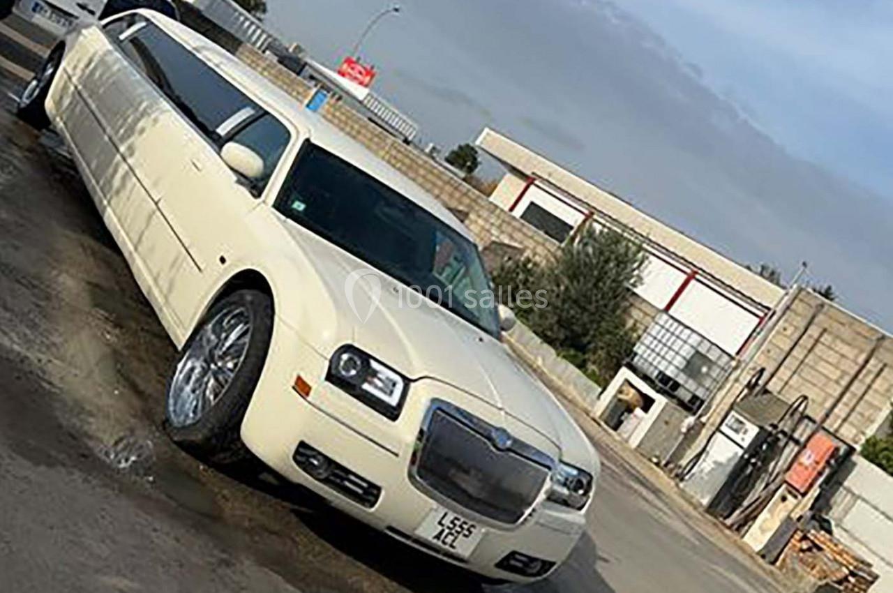 Limousine blanche stationnée sur un sol humide devant des bâtiments industriels sous un ciel partiellement nuageux.