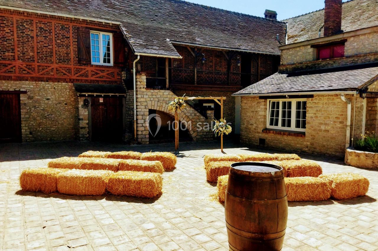 Cour pavée avec des bottes de paille disposées en cercle, entourée de bâtiments en pierre et en bois.