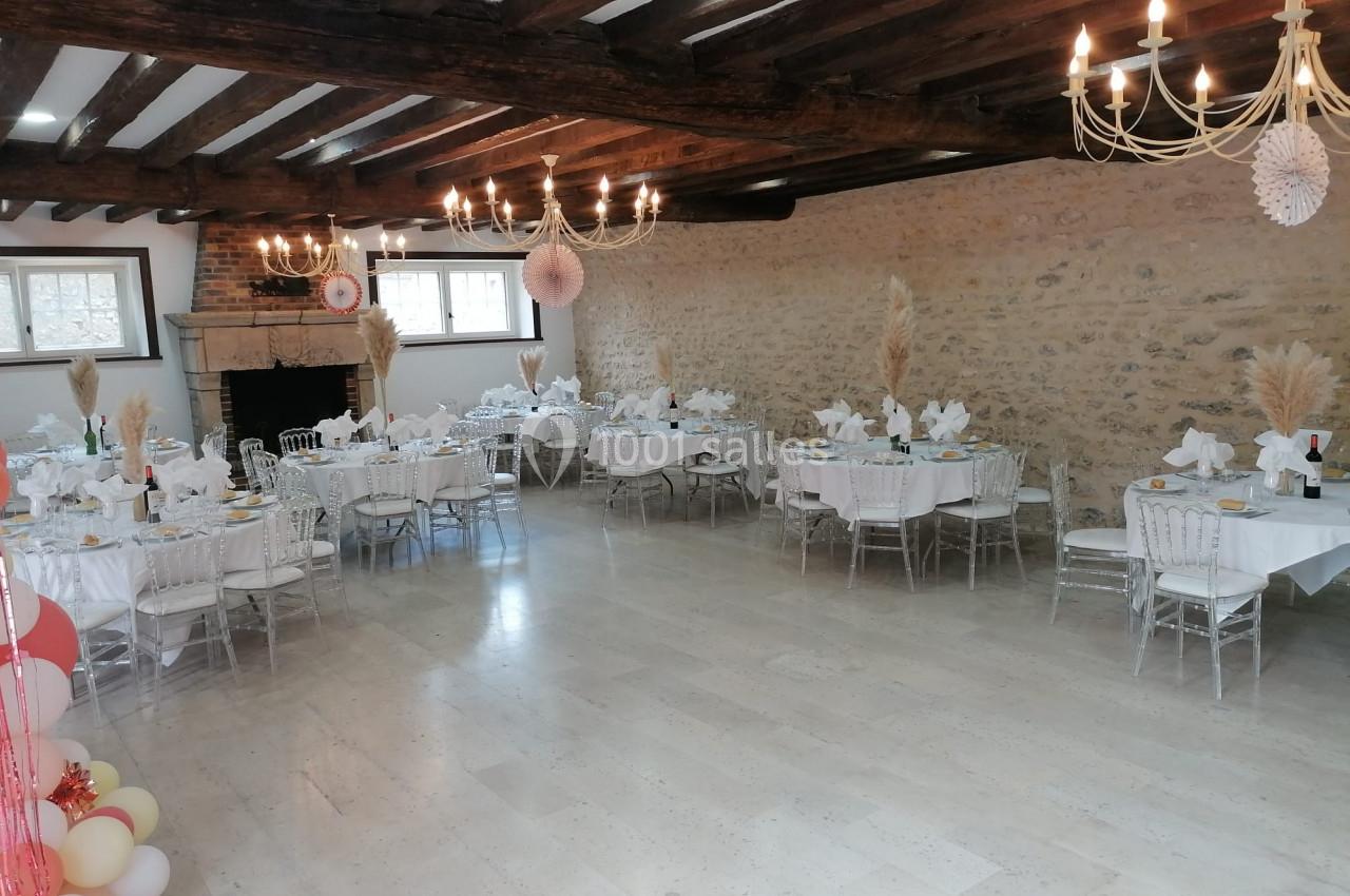 Salle de réception avec tables rondes dressées, décorations élégantes et poutres apparentes au plafond.