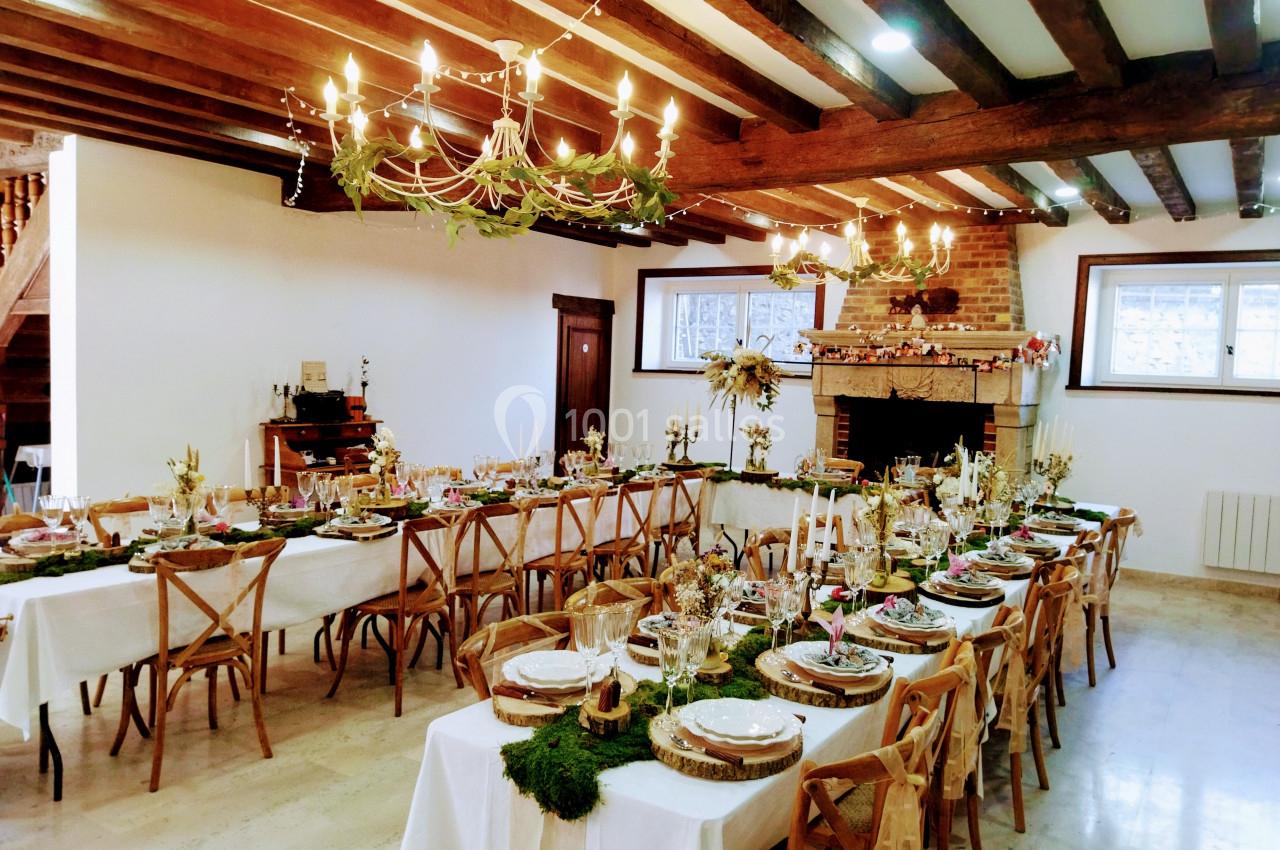 Salle décorée pour un repas festif, avec longues tables ornées de nappes blanches, bougies et éléments naturels.