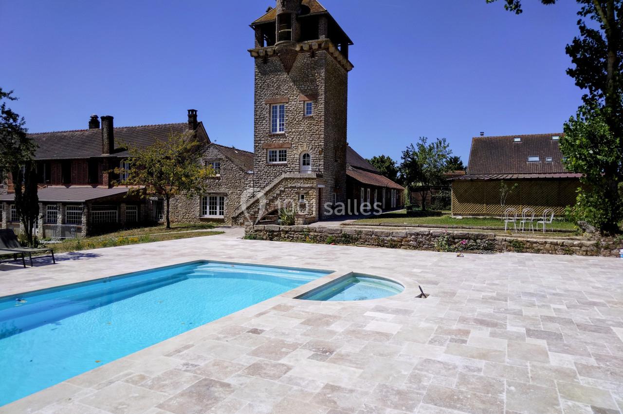 Grande piscine extérieure avec terrasse en pierre, vue sur une tour en pierre et des bâtiments traditionnels environnants.
