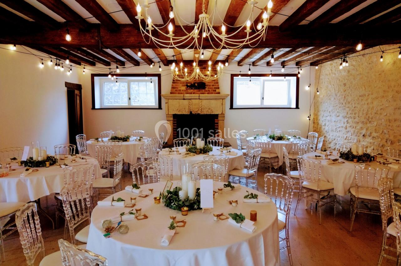 Salle de réception décorée avec des tables rondes, nappes blanches, chaises transparentes et guirlandes lumineuses…