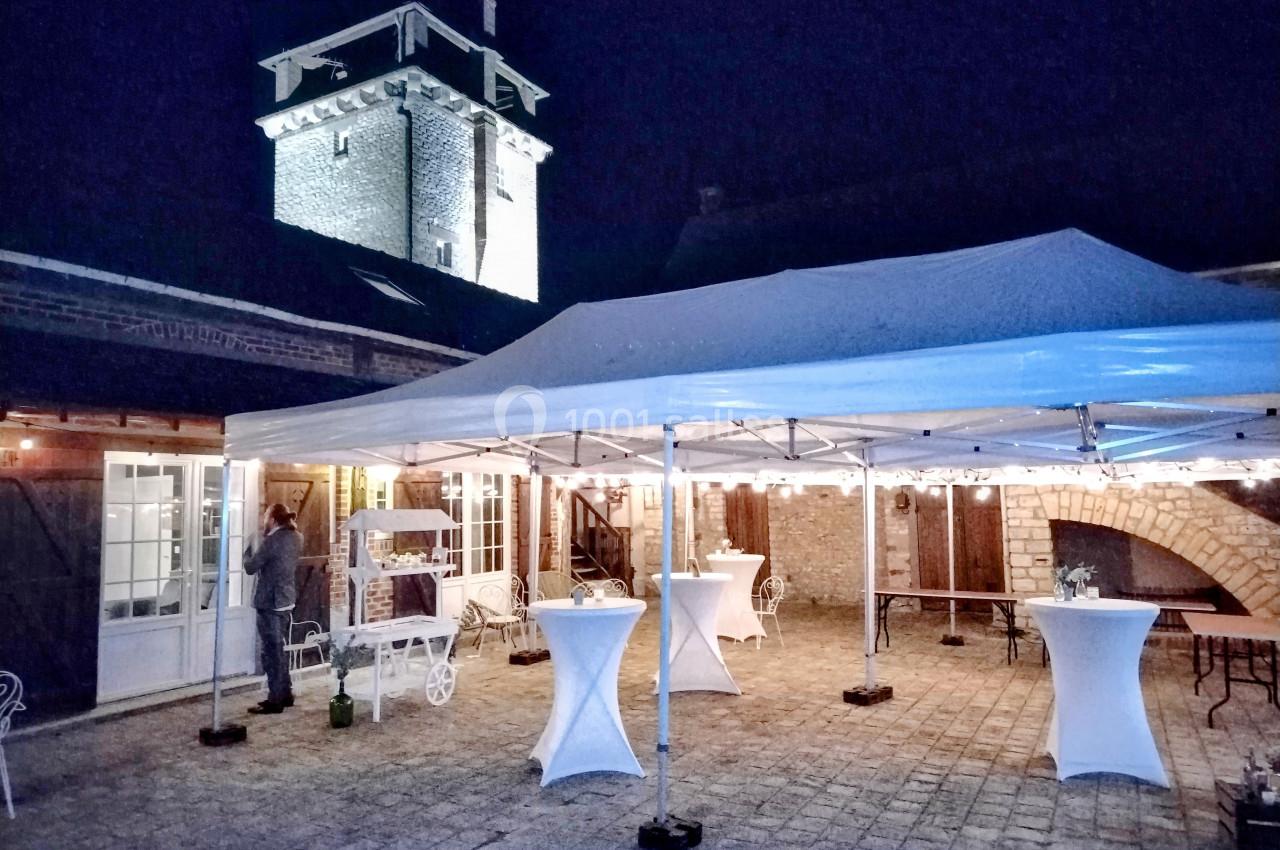 Cour éclairée de nuit avec une tente blanche, des tables hautes et un bâtiment en pierre avec une tour en arrière-plan.