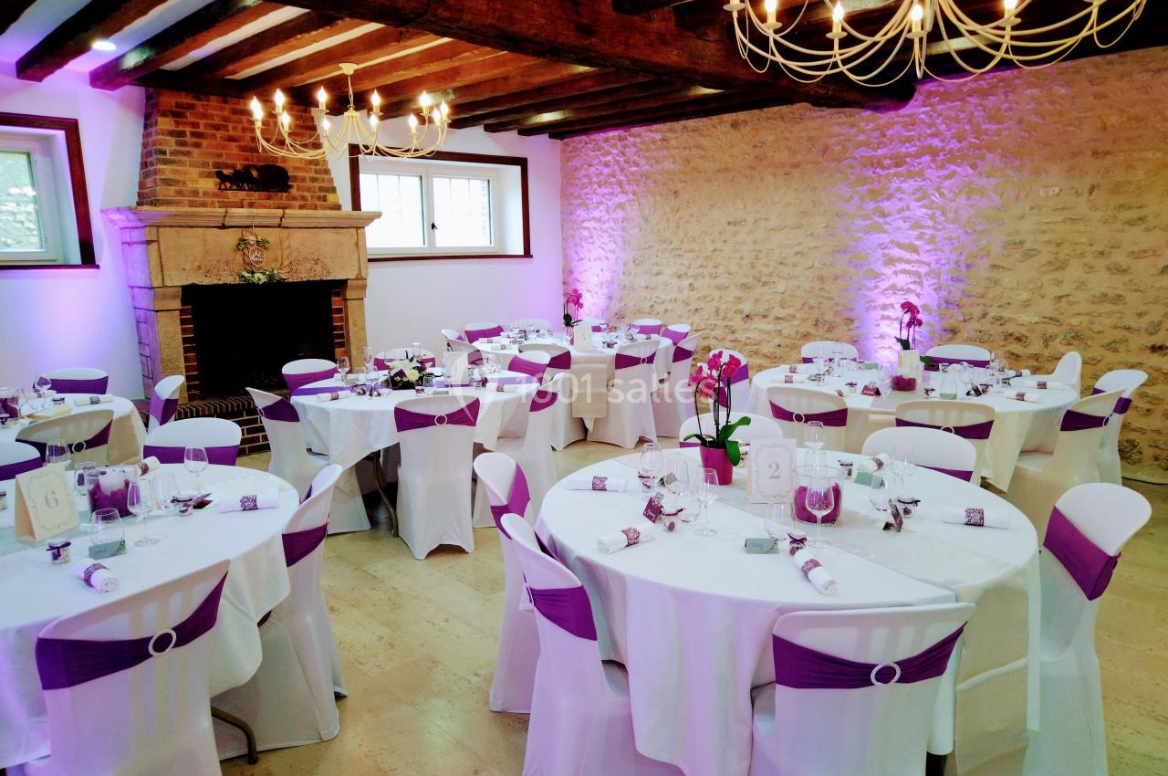 Salle de réception décorée avec des tables rondes, nappes blanches, chaises ornées de rubans violets et éclairage chaleureux.