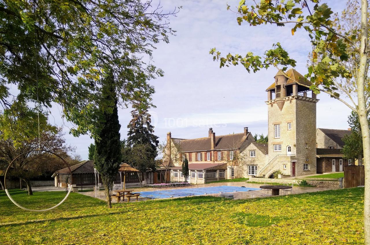 Grande maison en pierre avec une tour, entourée d'arbres et d'une pelouse couverte de feuilles, près d'une piscine vide.
