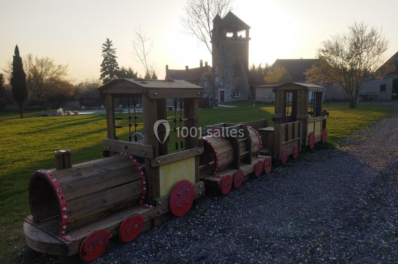 Train en bois pour enfants sur une pelouse, avec une tour en pierre et des arbres en arrière-plan au coucher du soleil.