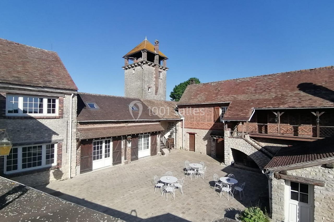 Cour intérieure pavée avec tables et chaises, entourée de bâtiments en pierre et d'une tour avec toit en bois.