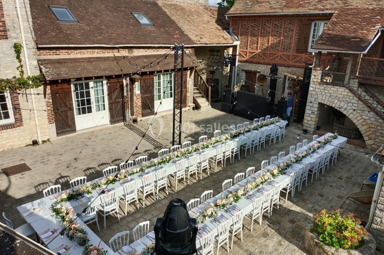 Tables de banquet décorées pour un événement en plein air dans une cour pavée entourée de bâtiments en pierre et en bois.