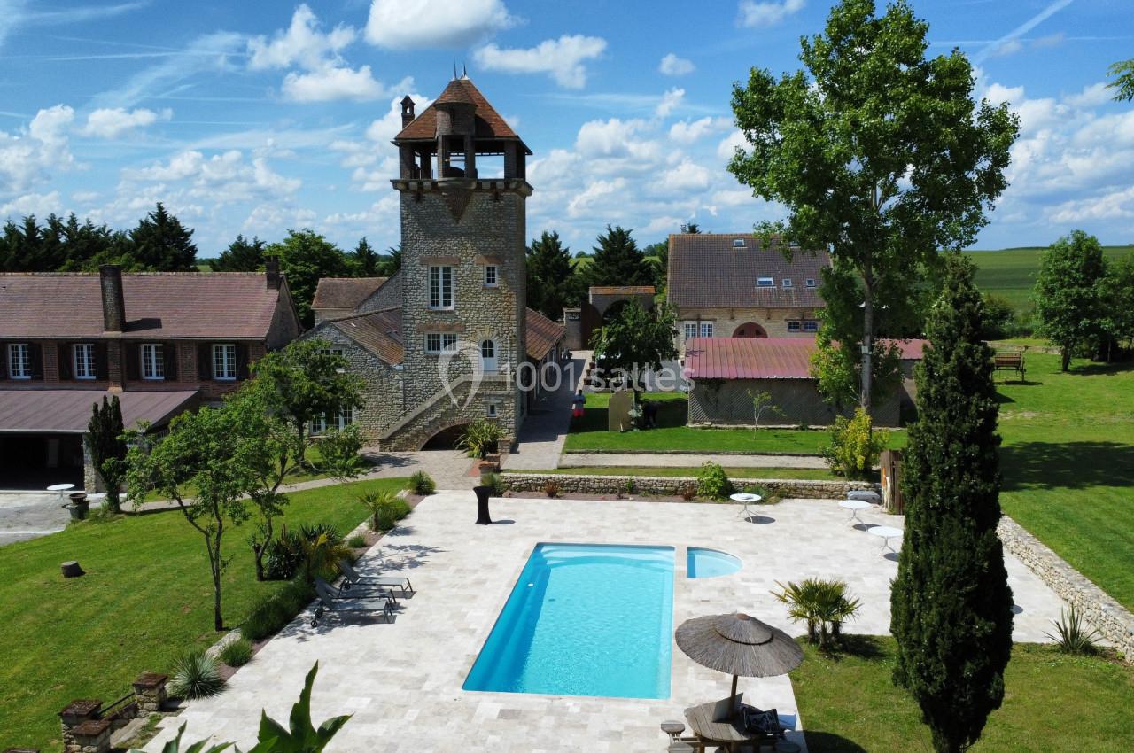 Vue d'un domaine avec une tour en pierre, une piscine entourée de verdure et des bâtiments annexes sous un ciel dégagé.