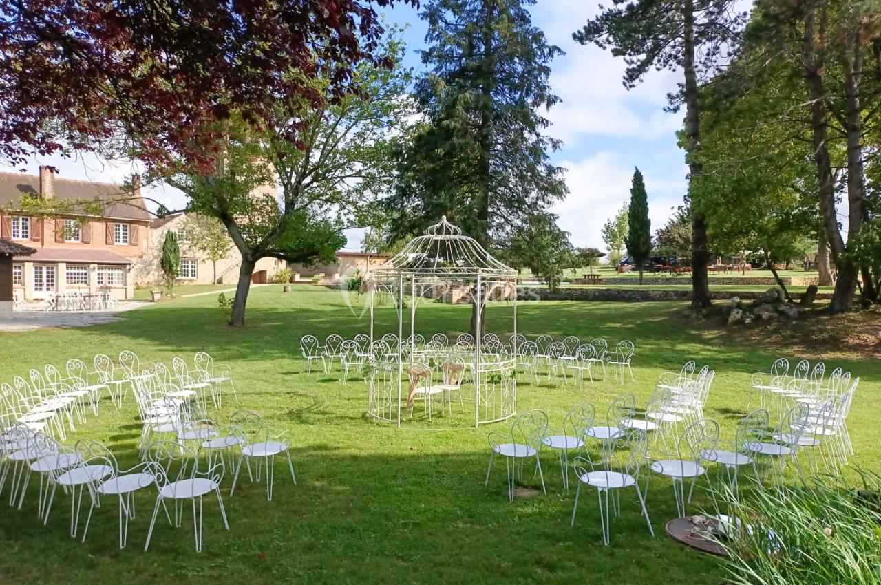 Chaises blanches disposées en cercle autour d'une pergola dans un jardin verdoyant près d'une maison en pierre.