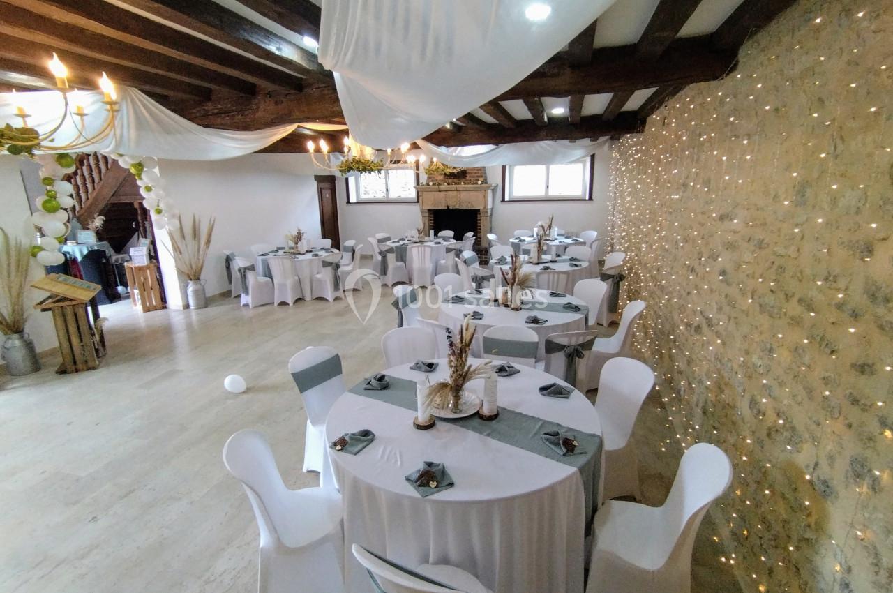 Salle de réception décorée avec des tables rondes, nappes blanches, chaises houssées et guirlandes lumineuses sur un mur en…