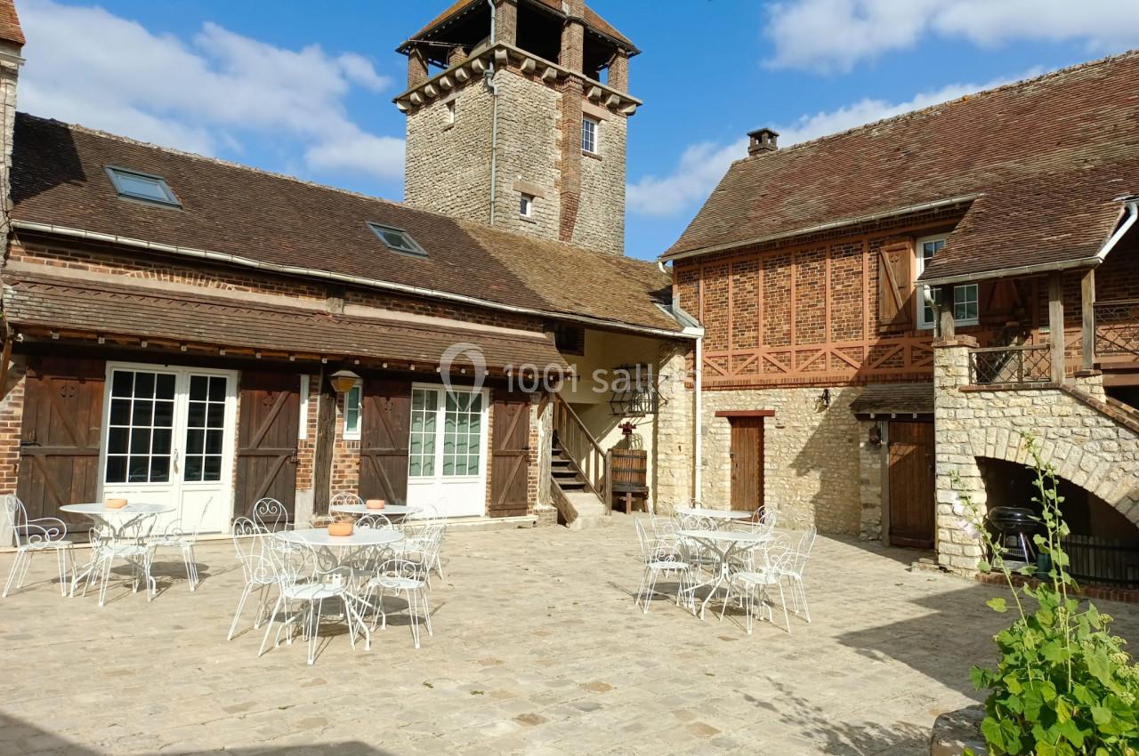 Cour pavée avec des tables et chaises en métal blanc, entourée de bâtiments anciens en pierre et bois sous un ciel bleu.