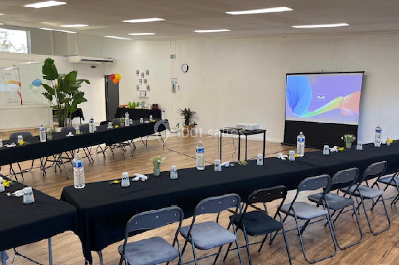Salle de réunion lumineuse avec deux longues tables noires, chaises pliantes, bouteilles d'eau et écran de projection allumé.
