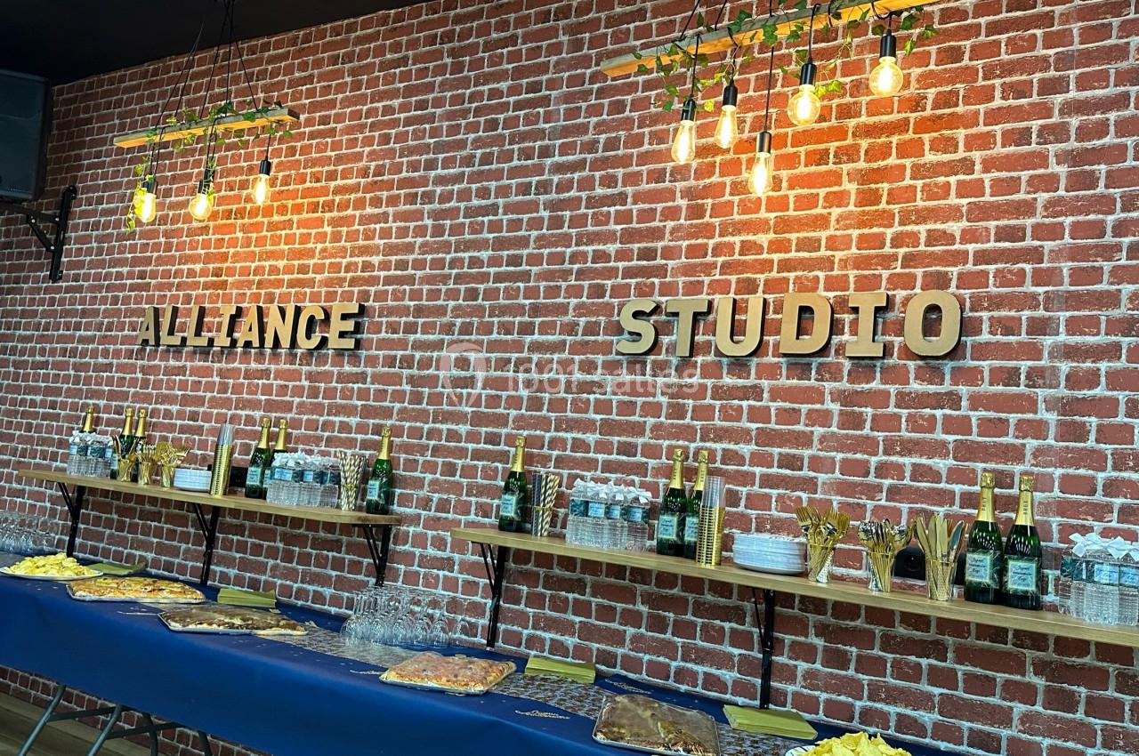Mur en briques rouges avec étagères contenant des bouteilles, verres et snacks, éclairé par des suspensions lumineuses.