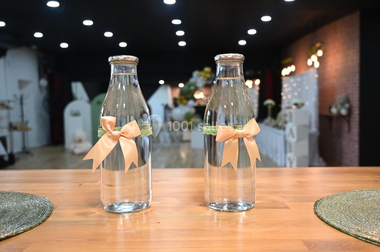 Deux bouteilles en verre décorées de rubans beige et vert posées sur une table en bois dans une salle éclairée.