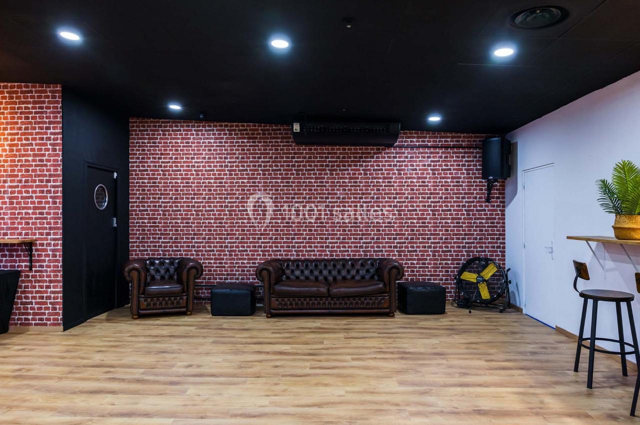 Salle avec mur en briques rouges, canapé en cuir marron, sol en bois clair et comptoir avec tabouret à droite.