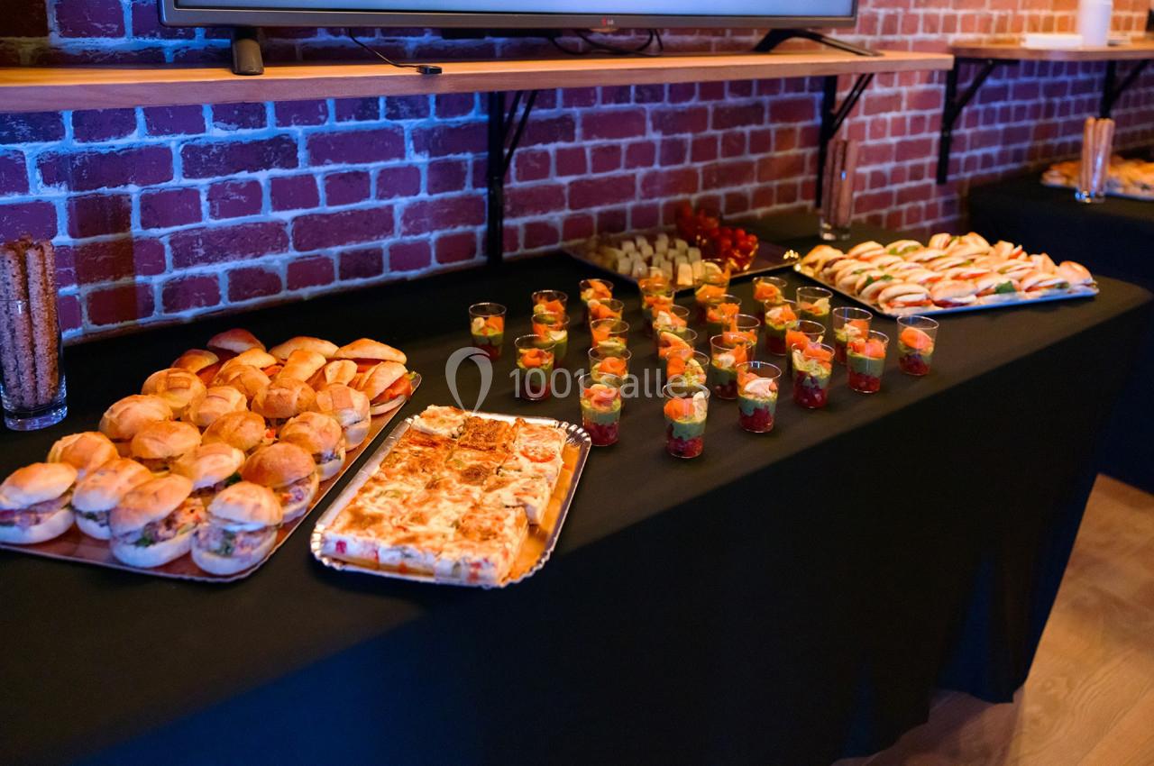 Buffet avec mini-sandwichs, verrines, parts de pizza et brochettes de fruits sur des nappes noires devant un mur en briques.
